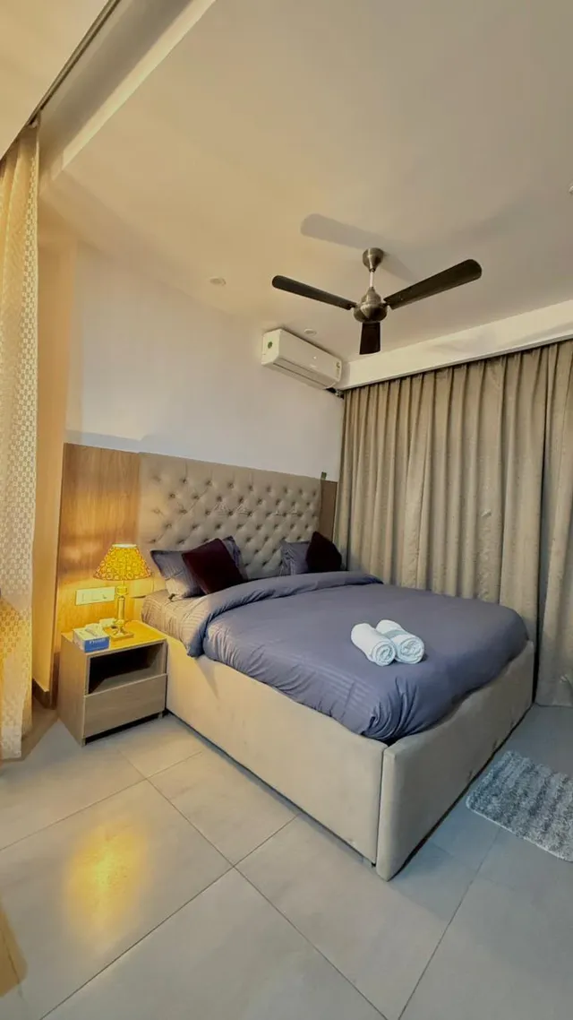 A bedroom with a padded headboard, grey bedding, warm bedside lighting and curtains.